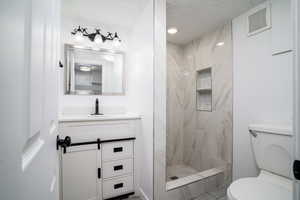 Bathroom featuring toilet, tiled shower, a textured ceiling, and vanity