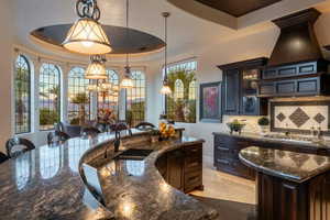 Kitchen with a sink, hanging light fixtures, a kitchen island with sink, dark stone countertops, and a tray ceiling