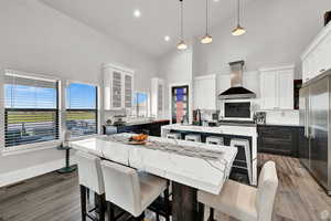 Kitchen with an island with sink, high end refrigerator, wall chimney exhaust hood, glass insert cabinets, and pendant lighting