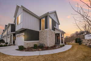 Property exterior at dusk with a lawn, driveway, stone siding, a garage, and board and batten siding