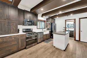 Kitchen featuring light wood finished floors, dark brown cabinets, appliances with stainless steel finishes, and beamed ceiling