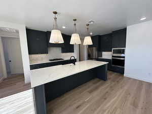 Kitchen featuring stainless steel appliances, dark cabinetry, an island with sink, and decorative light fixtures