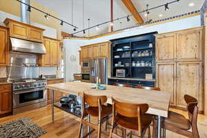 Kitchen featuring under cabinet range hood, a towering ceiling, appliances with stainless steel finishes, open shelves, and light wood finished floors