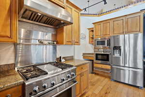 Kitchen with extractor fan, light wood-style flooring, appliances with stainless steel finishes, open shelves, and dark stone countertops