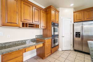 Kitchen with brown cabinets, light tile patterned floors, stainless steel appliances, built in study area, and dark stone counters