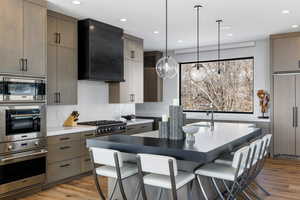 Kitchen with pendant lighting, dark countertops, modern cabinets, built in appliances, and wall chimney exhaust hood