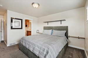 Bedroom with baseboards, visible vents, dark colored carpet, and ensuite bathroom