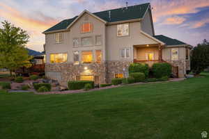 Back of house at dusk with stone siding, a yard, roof with shingles, a wooden deck, and stucco siding