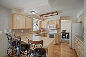 Kitchen with light brown cabinetry, a sink, white appliances, a peninsula, and a kitchen bar