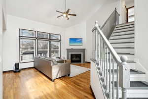 Living area with ceiling fan, high vaulted ceiling, wood finished floors, a fireplace with flush hearth, and stairway