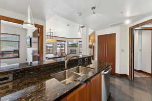 Kitchen featuring brown cabinetry, a sink, dishwasher, decorative light fixtures, and dark stone counters