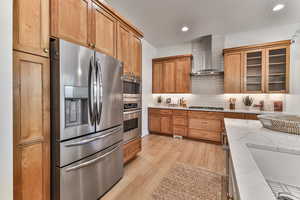 Kitchen with light stone counters, wall chimney range hood, light hardwood / wood-style floors, stainless steel appliances, and tasteful backsplash