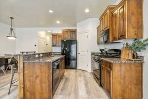 Kitchen with a breakfast bar area, a center island with sink, granite countertop, brown cabinets, black appliances, and decorative light fixtures