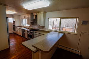 Kitchen featuring a baseboard heating unit, appliances with stainless steel finishes, a sink, a peninsula, and wall chimney exhaust hood