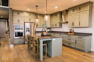 Kitchen with dark countertops, an island with sink, wall chimney exhaust hood, appliances with stainless steel finishes, and pendant lighting