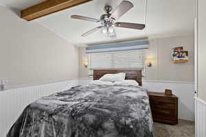 Carpeted bedroom with vaulted ceiling with beams, a wainscoted wall, and a ceiling fan