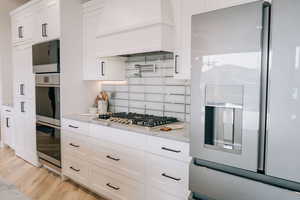 Kitchen with tasteful backsplash, white cabinets, light stone counters, custom exhaust hood, and stainless steel appliances