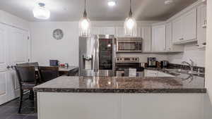 Kitchen with stainless steel appliances, a sink, white cabinets, hanging light fixtures, and dark stone counters