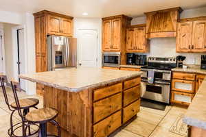 Kitchen featuring a kitchen island, light countertops, appliances with stainless steel finishes, custom exhaust hood, and brown cabinetry