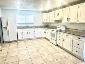 Kitchen with under cabinet range hood, white appliances, a sink, white cabinets, and dark stone counters