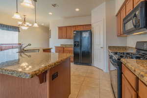 Kitchen featuring hanging light fixtures, brown cabinetry, a kitchen island with sink, a sink, and black appliances