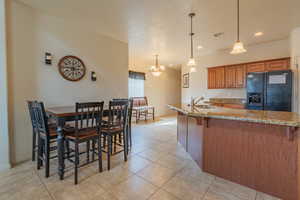 Kitchen featuring light stone counters, visible vents, hanging light fixtures, black fridge, and brown cabinetry