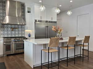 Kitchen with high-quality appliances, white cabinetry, hanging light fixtures, wall chimney exhaust hood, and glass insert cabinets