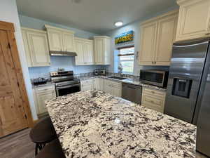 Kitchen with cream cabinets, appliances with stainless steel finishes, light stone counters, and a sink