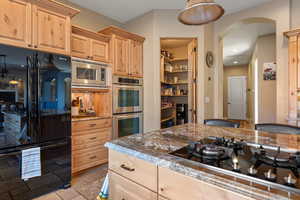 Kitchen with arched walkways, dark stone countertops, stone tile flooring, black appliances, and light brown cabinets