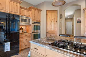 Kitchen with stone tile floors, arched walkways, dark stone countertops, light brown cabinetry, and black appliances