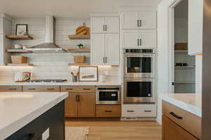 Kitchen featuring stainless steel appliances, light wood-type flooring, wall chimney range hood, white cabinetry, and open shelves