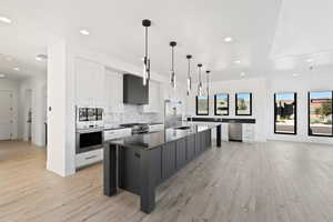 Kitchen with built in appliances, a kitchen island with sink, white cabinetry, dark countertops, and decorative light fixtures