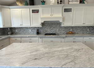 Kitchen with light stone counters, stainless steel gas stovetop, backsplash, glass insert cabinets, and white cabinets