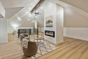 Living room featuring baseboards, a glass covered fireplace, lofted ceiling with beams, light wood-style flooring, and ceiling fan