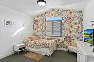 Bedroom featuring wallpapered walls, baseboards, vaulted ceiling, and dark colored carpet