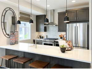 Kitchen featuring stainless steel appliances, dark brown cabinets, light countertops, wall chimney range hood, and a kitchen bar