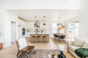 Kitchen featuring pendant lighting, light countertops, a kitchen island with sink, white cabinetry, and wall chimney range hood