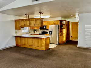 Kitchen featuring light carpet, a peninsula, light countertops, appliances with stainless steel finishes, and brown cabinetry