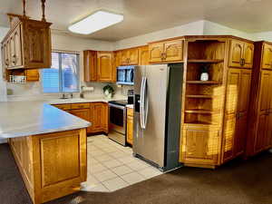 Kitchen featuring appliances with stainless steel finishes, brown cabinets, a peninsula, light countertops, and open shelves