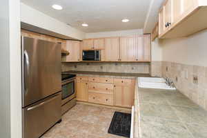 Kitchen featuring tile counters, appliances with stainless steel finishes, a textured ceiling, light brown cabinets, and a sink