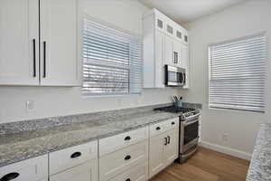 Kitchen with light wood-style flooring, glass insert cabinets, appliances with stainless steel finishes, light stone countertops, and white cabinetry