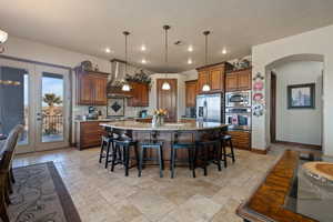 Kitchen with pendant lighting, stone tile flooring, appliances with stainless steel finishes, a large island with sink, and wall chimney exhaust hood