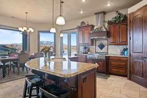 Kitchen featuring decorative light fixtures, a sink, wall chimney range hood, an island with sink, and stone tile flooring