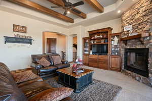 Living area featuring arched walkways, ceiling fan, beamed ceiling, a stone fireplace, and carpet floors