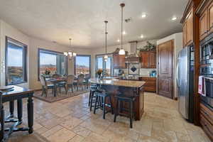 Kitchen featuring pendant lighting, stone tile flooring, appliances with stainless steel finishes, a kitchen island, and wall chimney exhaust hood