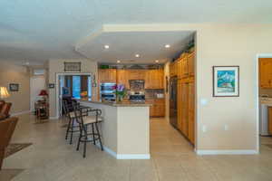 Kitchen with light tile patterned floors, stainless steel appliances, a kitchen island with sink, dark stone countertops, and a kitchen bar