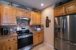 Kitchen featuring light tile patterned floors, dark granite countertops, stainless steel appliances, under cabinet range hood, and tile backsplash