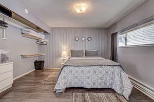 Bedroom with a textured ceiling, wooden walls, a baseboard heating unit, wood finished floors, and baseboards