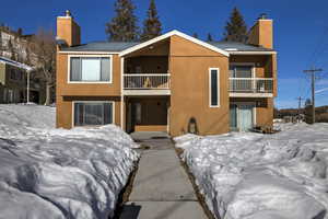 Snow covered back of property featuring metal roof, a chimney, a balcony, and stucco siding