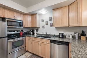 Kitchen featuring light tile patterned floors, dark stone counters, stainless steel appliances, light brown cabinetry, and a sink